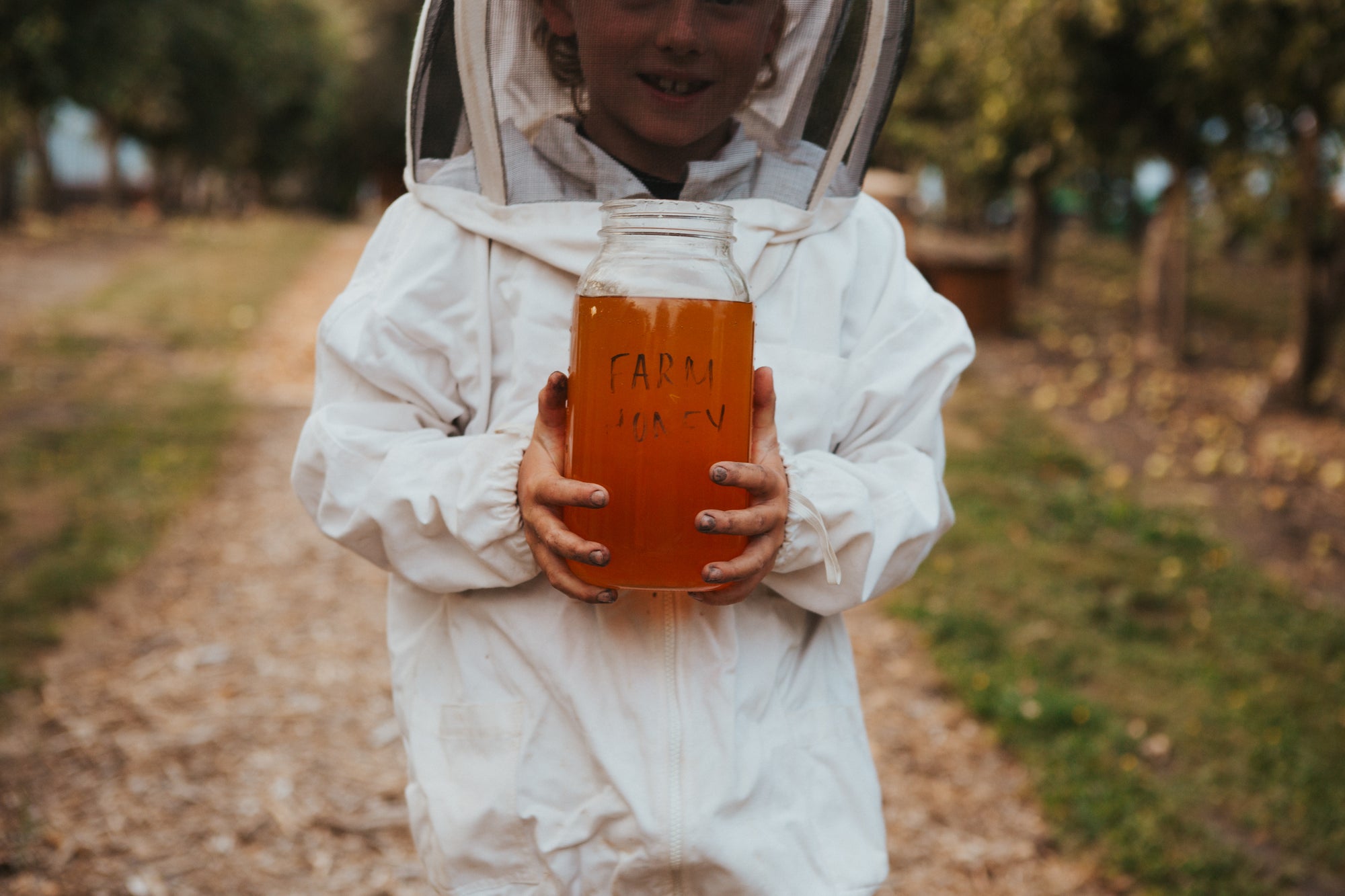 A child wearing a white beekeeping suit with a clear face shield holds a glass jar filled with amber honey outdoors on a dirt path.